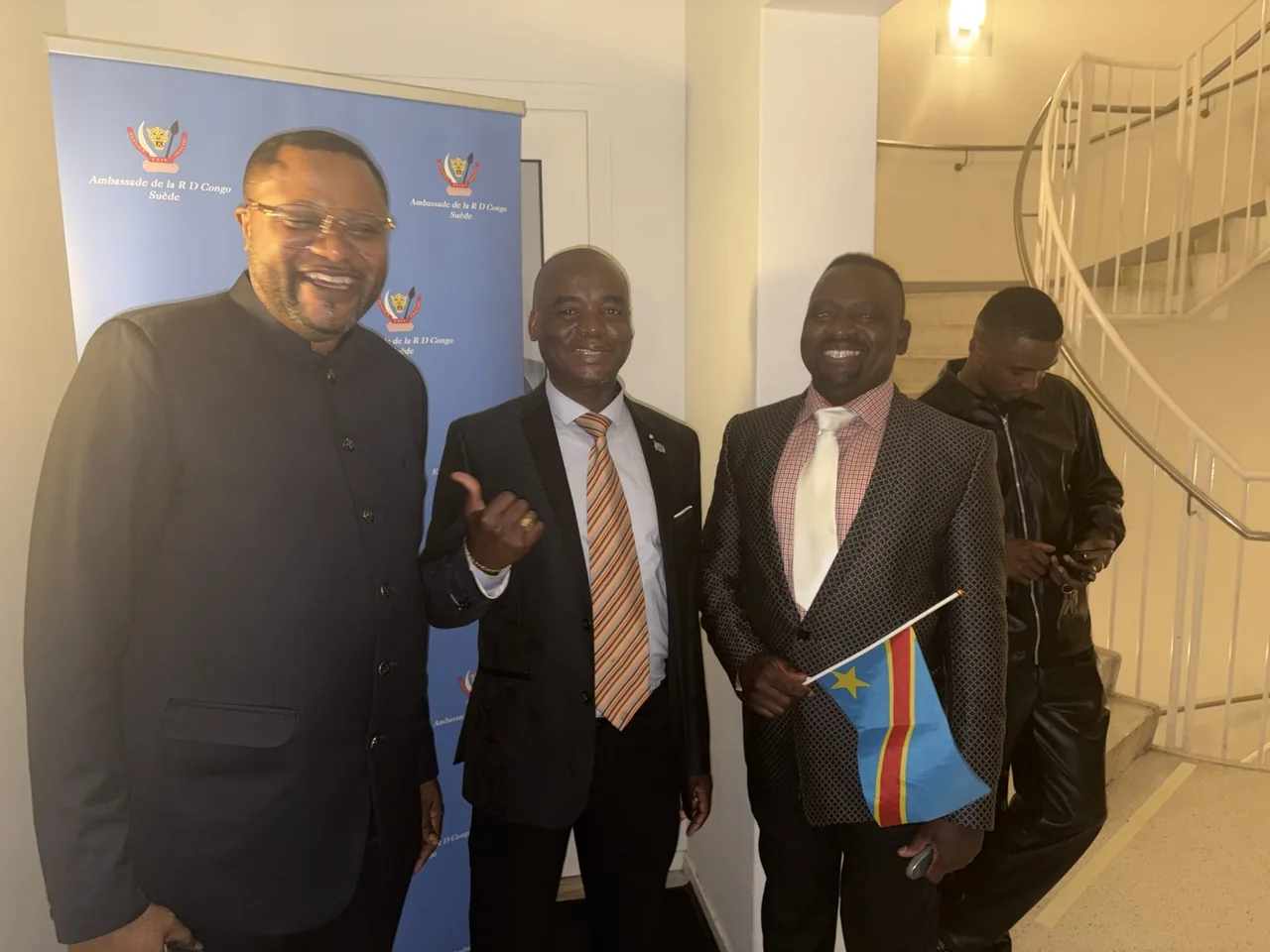 Groupe d'hommes souriants posant ensemble près d'un stand de l'Ambassade de la RD Congo en Suède, l'un tenant un petit drapeau congolais.